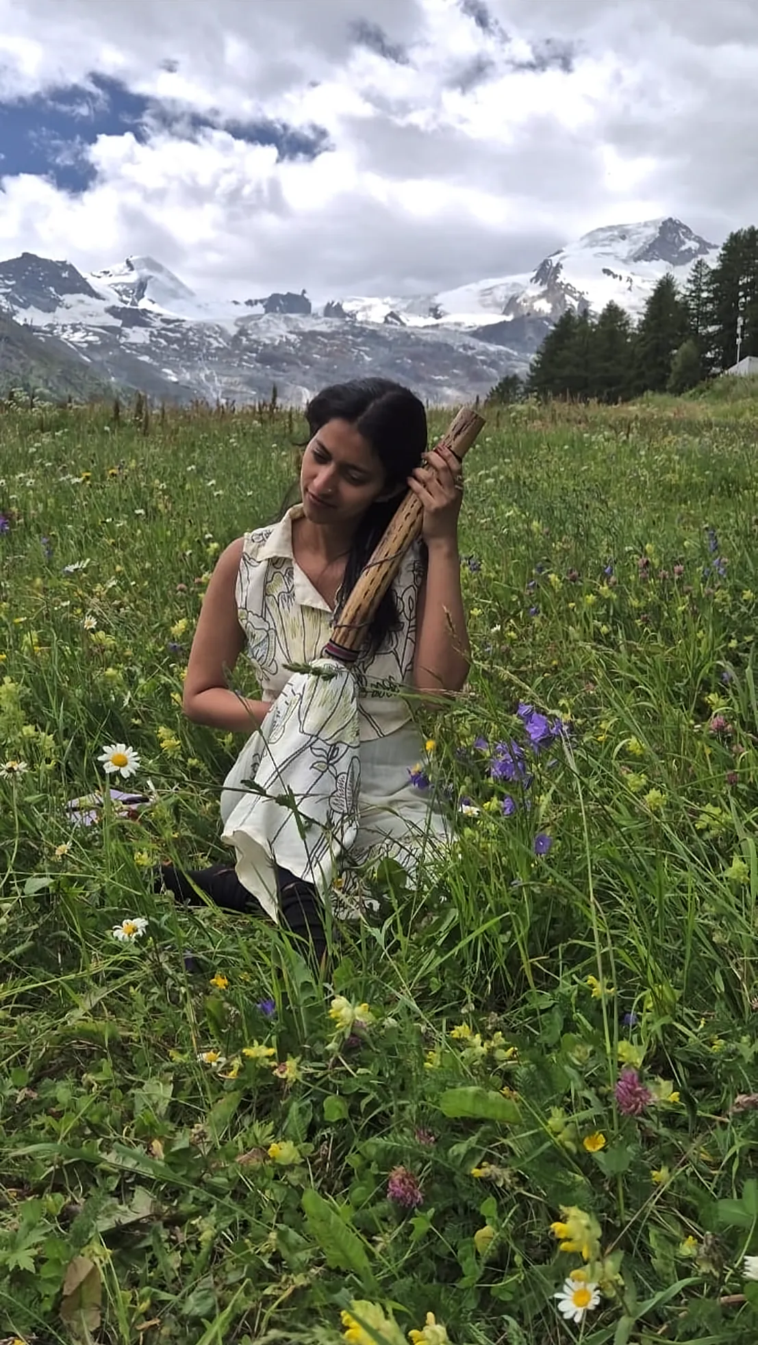 Ritty kneeling in a wildflower meadow below snowy mountains, holding a rain stick to her ear
