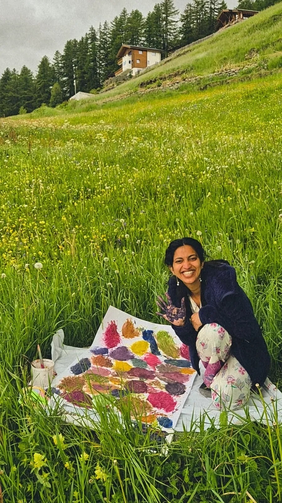 Ritty kneeling beside her colourful painting in a mountain meadow